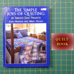 May include: A blue book titled "The Simple Joys of Quilting: 30 Timeless Quilt Projects" by Joan Hanson and Mary Hickey. The cover features a photograph of a bed with a colorful quilt.