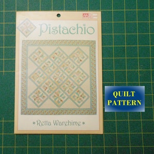May include: A quilt pattern titled "Pistachio" by Rella Warehime. The pattern features a quilt design with a light blue and cream color scheme. The pattern is printed on a white card with a blue border.