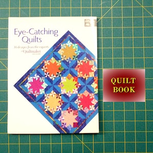 May include: A book titled "Eye-Catching Quilts" with a colorful quilt design on the cover. The book is on a green cutting mat with a red label that says "QUILT BOOK".