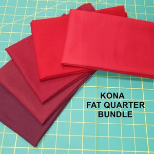 May include: A bundle of Kona cotton fat quarters in shades of red, from deep burgundy to bright red. The fabric is neatly stacked on a green cutting mat, with the text "KONA FAT QUARTER BUNDLE" in white.
