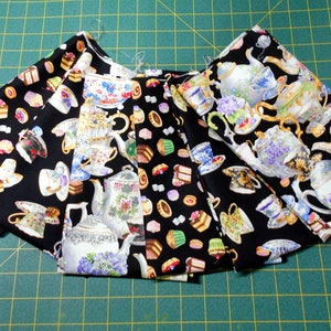 May include: A collection of six fabric squares with different patterns featuring teacups, teapots, and pastries on a black background. The fabric squares are arranged in a semi-circle on a green cutting mat.