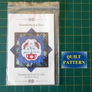 May include: A white paper quilt pattern with a blue and red design of two lovebirds in a tree with a red cup underneath. The title of the pattern is "Lovebirds in a Tree" and the text below reads "Pennsylvania Dutch Hex Signs By: Bonnie Kaster".