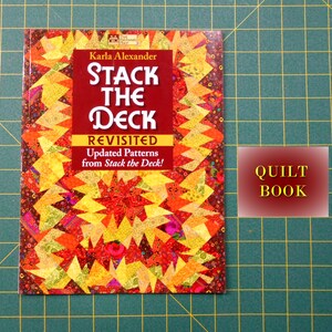 May include: A book titled "Stack the Deck Revisited" by Karla Alexander. The cover features a colorful quilt pattern with red, yellow, and orange stars. The book is a quilting guide with updated patterns from the original "Stack the Deck" book. The text "QUILT BOOK" is visible on a red background.