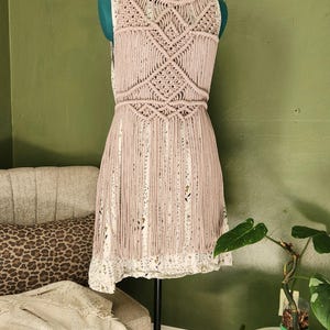 May include: A sleeveless dress featuring a light mauve macrame overlay with intricate knotted geometric patterns and long fringe detailing. The dress underneath is white with a subtle floral print, featuring a round neckline and a straight hem.