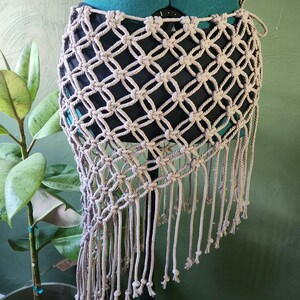 May include: A beige macrame skirt featuring a diamond-shaped knotted pattern and long fringe trim along the bottom edge. The skirt is displayed on a mannequin over a solid black base layer, showcasing the intricate cord work and open weave design.