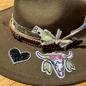 May include: A green felt cowboy hat with a black heart-shaped rhinestone embellishment, a floral skull patch, and a feather accent. The hat also has a brown leather band with a white card that says "8" on it.