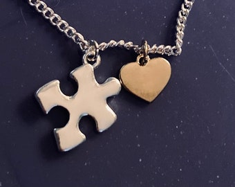 May include: A silver-toned metal chain necklace featuring two charms: a white puzzle piece and a small, smooth gold-toned heart. The charms are attached to the chain by small metal jump rings, creating a simple and distinct jewelry piece.