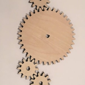 May include: Four wooden gears interlocked together. The largest gear is in the center and the three smaller gears are around it. The gears are all made of light brown wood and have black outlines.