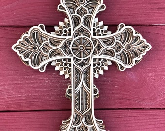 May include: A wooden cross with intricate laser-cut designs. The cross is made of multiple layers of wood, creating a 3D effect. The design features a central circle with a smaller circle inside, surrounded by floral patterns.