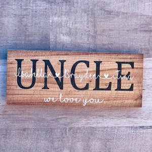 May include: Wooden sign with the word "UNCLE" in black lettering. The sign also includes the names "Isabella", "Brayden", and "Tina" in white lettering with a heart symbol between each name. The sign reads "we love you" in white lettering.