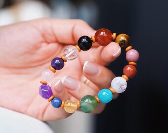 Puede incluir: Una pulsera de cuentas coloridas con una variedad de piedras preciosas en tonos morados, azules, verdes, amarillos, naranjas y negros. Las cuentas están ensartadas con elásticos y pequeños espaciadores de madera. La pulsera se sostiene en una mano.
