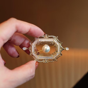 Puede incluir: Broche dorado con una perla central, engastada en un marco rectangular. El marco está adornado con pequeños detalles brillantes y delicados detalles de filigrana. El broche tiene una estética vintage.