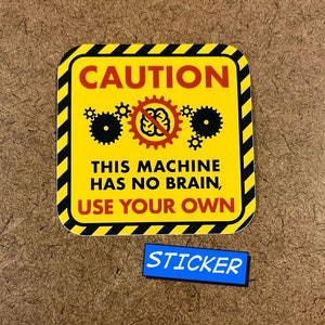 May include: A yellow sticker with a black border and the word "CAUTION" in red. The sticker features a graphic of gears and a brain with a line through it. The text "THIS MACHINE HAS NO BRAIN, USE YOUR OWN" is below the graphic. A small blue sticker with the word "STICKER" is below.