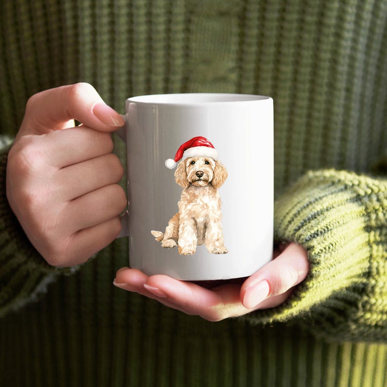Pode incluir: Caneca de cer&acirc;mica branca com uma ilustra&ccedil;&atilde;o em aquarela de um golden doodle usando um gorro de Papai Noel vermelho. O c&atilde;o est&aacute; sentado, com uma pelagem fofa e uma express&atilde;o doce. A caneca &eacute; segurada por uma pessoa vestindo um su&eacute;ter verde.