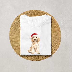 Pode incluir: Camiseta branca com uma ilustra&ccedil;&atilde;o em aquarela de um c&atilde;o golden doodle usando um chap&eacute;u de Papai Noel. O c&atilde;o tem uma pelagem marrom clara e um chap&eacute;u vermelho e branco. A camiseta &eacute; dobrada em uma superf&iacute;cie circular tecida.