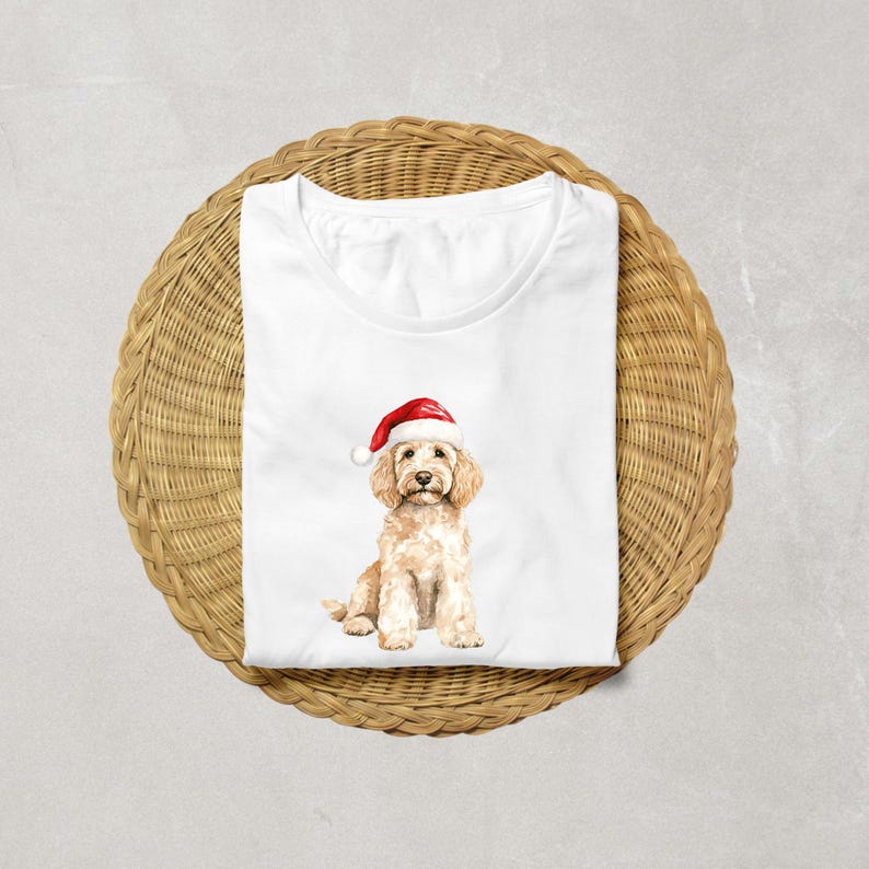 Pode incluir: Camiseta branca com uma ilustra&ccedil;&atilde;o em aquarela de um c&atilde;o golden doodle usando um chap&eacute;u de Papai Noel. O c&atilde;o tem uma pelagem marrom clara e um chap&eacute;u vermelho e branco. A camiseta &eacute; dobrada em uma superf&iacute;cie circular tecida.