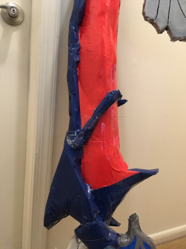 May include: A red and blue cardboard prop, possibly a weapon or a part of a costume. The prop is made of cardboard and covered in blue and red tape.