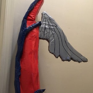 May include: A red and blue cardboard sculpture of an angel with a single wing. The wing is made of gray cardboard and has a detailed design.