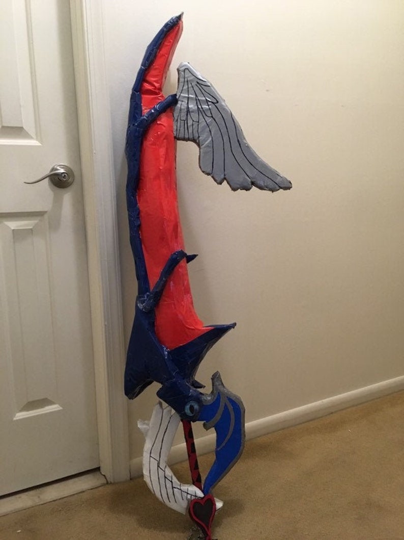May include: A handmade cardboard replica of a video game weapon. The weapon is red, blue, and gray with a heart-shaped handle.