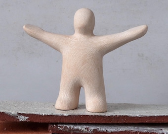 May include: A beige ceramic figurine of a person with arms outstretched. The sculpture has a minimalist design and stands on a gray and red surface. The figure's arms are extended, giving the impression of welcoming or embracing.
