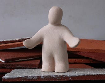 May include: A light beige ceramic figurine of a person with outstretched arms. The figure stands on a gray and red surface, with red rectangular tiles in the background. The figurine has a smooth, matte finish.