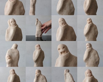 May include: A collection of beige ceramic sculptures depicting various facial expressions. The sculptures are wrapped in a hooded cloak, with each piece showcasing a different emotion, including joy, sadness, and surprise. The sculptures are displayed against a neutral background.