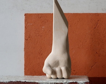 May include: A beige sculpture of a clenched fist with a sharp, triangular extension. The artwork is set against a backdrop of a red brick and a white surface, creating a contrast in textures and colors. The sculpture is on a dark surface.