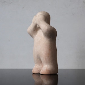 Può includere: Una scultura astratta beige di una figura con le mani che coprono le orecchie. La scultura ha una forma liscia e arrotondata e sta in piedi. Le gambe della figura sono leggermente divaricate e il design è minimalista.