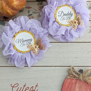 May include: Two purple and white lace flower pins with gold glitter circles that say "Mommy to Be" and "Daddy to Be". Each pin has a gold butterfly with a rhinestone on it. The pins are on a white wooden background with a small orange pumpkin.
