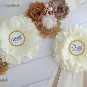 Puede incluir: Dos fajas de flores blancas con ribetes dorados y texto negro. Una faja dice "Mommy To Be" y la otra dice "Daddy To Be". Las fajas est&aacute;n decoradas con flores de tela marr&oacute;n y dorado.