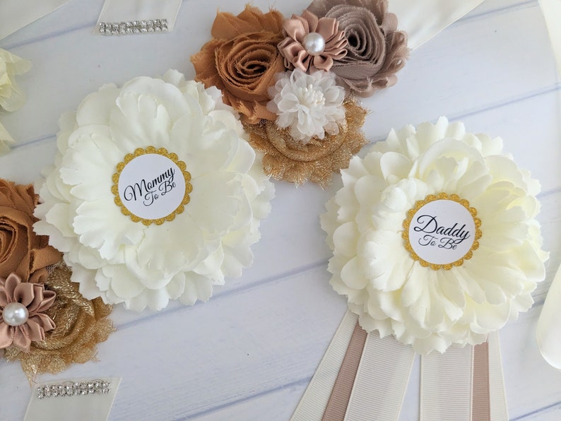 Puede incluir: Dos fajas de flores blancas con ribetes dorados y texto negro. Una faja dice "Mommy To Be" y la otra dice "Daddy To Be". Las fajas est&aacute;n decoradas con flores de tela marr&oacute;n y dorado.