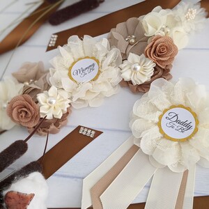 Puede incluir: Dos fajas con cintas marrones y beige y flores de encaje blanco. Una faja tiene un c&iacute;rculo blanco con el texto "Mommy To Be" y la otra tiene un c&iacute;rculo blanco con el texto "Daddy To Be".