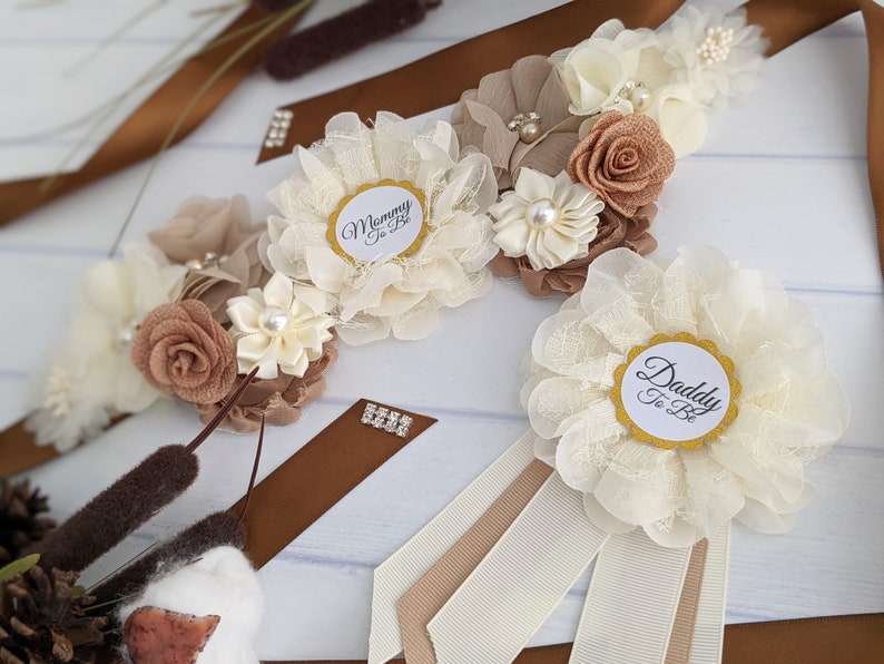 Puede incluir: Dos fajas con cintas marrones y beige y flores de encaje blanco. Una faja tiene un c&iacute;rculo blanco con el texto "Mommy To Be" y la otra tiene un c&iacute;rculo blanco con el texto "Daddy To Be".