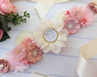 May include: A pink and white floral sash with a gold "Mommy To Be" circle in the center. The sash is decorated with rhinestones and pearls.
