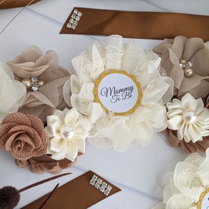 Puede incluir: Una faja floral blanca y marr&oacute;n con un c&iacute;rculo dorado que dice "Mommy To Be".