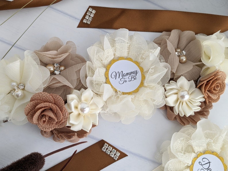 Puede incluir: Una faja floral blanca y marr&oacute;n con un c&iacute;rculo dorado que dice "Mommy To Be".