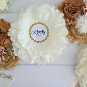 Puede incluir: Una faja de sat&eacute;n blanco con una flor blanca grande y una flor beige m&aacute;s peque&ntilde;a con un acento de perla. La faja tiene una tela de brillo dorado y un c&iacute;rculo blanco con el texto "Mommy To Be" en negro.