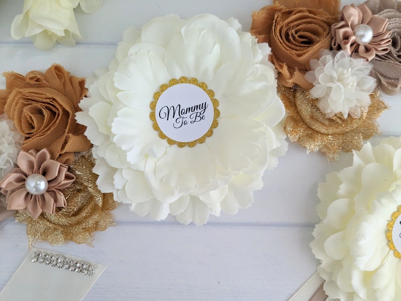 Puede incluir: Una faja de sat&eacute;n blanco con una flor blanca grande y una flor beige m&aacute;s peque&ntilde;a con un acento de perla. La faja tiene una tela de brillo dorado y un c&iacute;rculo blanco con el texto "Mommy To Be" en negro.