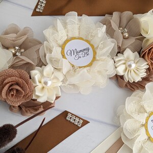 Puede incluir: Una faja marr&oacute;n y marfil con una gran flor marfil y una flor marr&oacute;n m&aacute;s peque&ntilde;a. La faja tiene un c&iacute;rculo dorado con el texto "Mommy To Be" y un c&iacute;rculo dorado con el texto "Daddy To Be".