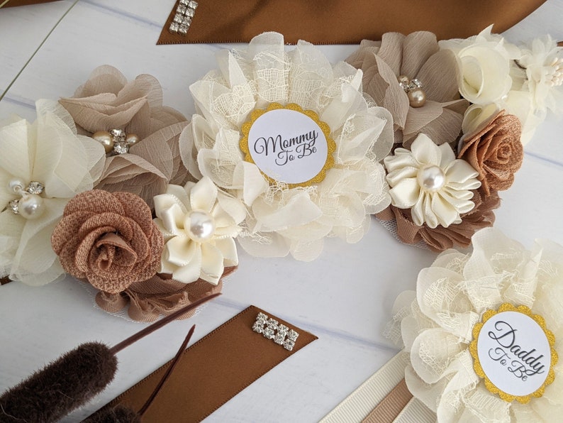 Puede incluir: Una faja marr&oacute;n y marfil con una gran flor marfil y una flor marr&oacute;n m&aacute;s peque&ntilde;a. La faja tiene un c&iacute;rculo dorado con el texto "Mommy To Be" y un c&iacute;rculo dorado con el texto "Daddy To Be".