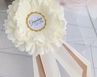 May include: White flower with a gold circle that says "Grandma To Be" and a white ribbon with a brown ribbon underneath.
