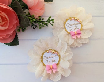 May include: Two white lace flower pins with gold glitter and pink bows. The pins say "Grandma To Be" and "Grandpa To Be".