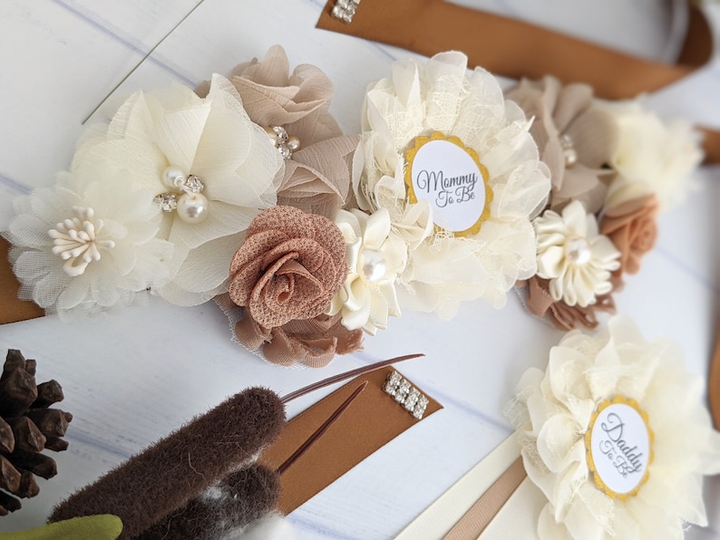 Puede incluir: Una faja marr&oacute;n con un arreglo floral blanco y marr&oacute;n. La faja tiene un c&iacute;rculo dorado con el texto "Mommy To Be" y un c&iacute;rculo dorado con el texto "Daddy To Be".