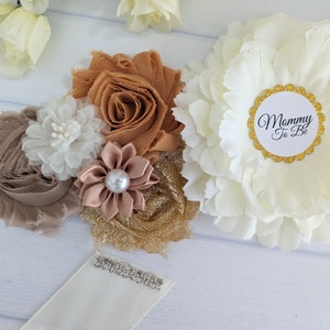 Puede incluir: Una faja de sat&eacute;n blanco con un ribete de purpurina dorada y una gran flor blanca con un c&iacute;rculo dorado que dice "Mommy To Be". La faja est&aacute; decorada con flores de tela marr&oacute;n, dorado y blanco.