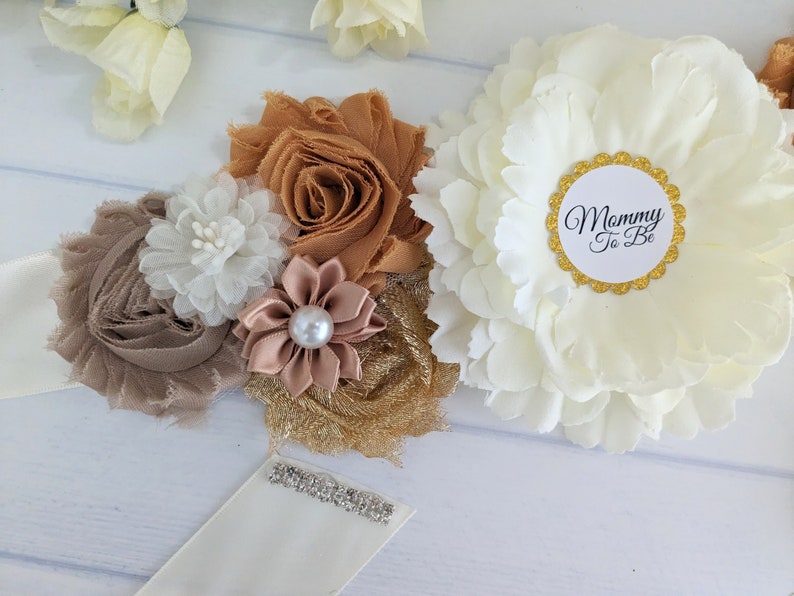 Puede incluir: Una faja de sat&eacute;n blanco con un ribete de purpurina dorada y una gran flor blanca con un c&iacute;rculo dorado que dice "Mommy To Be". La faja est&aacute; decorada con flores de tela marr&oacute;n, dorado y blanco.
