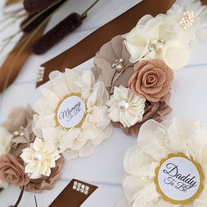Puede incluir: Una faja floral blanca y marr&oacute;n con el texto "Mommy to Be" y "Daddy to Be" en dos c&iacute;rculos blancos separados. La faja est&aacute; decorada con flores blancas y marrones, pedrer&iacute;a y purpurina dorada.