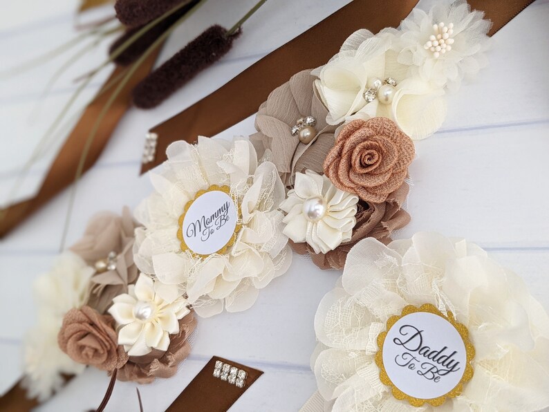 Puede incluir: Una faja floral blanca y marr&oacute;n con el texto "Mommy to Be" y "Daddy to Be" en dos c&iacute;rculos blancos separados. La faja est&aacute; decorada con flores blancas y marrones, pedrer&iacute;a y purpurina dorada.