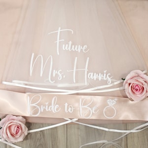 May include: White tulle bridal veil with a white satin sash that says "Bride to Be" with a diamond ring icon. The veil also has the text "Future Mrs. Harris" written on it.