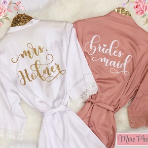 May include: Two white satin robes with lace trim. One robe says "mrs. Holmer" in gold glitter lettering. The other robe says "bridesmaid" in white lettering.