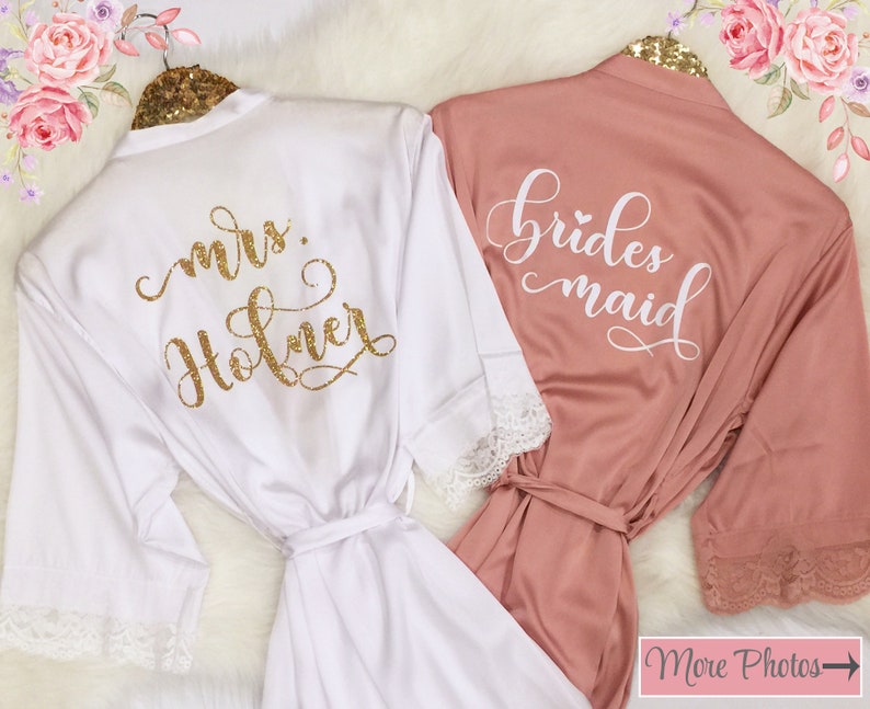 May include: Two white satin robes with lace trim. One robe says "mrs. Holmer" in gold glitter lettering. The other robe says "bridesmaid" in white lettering.